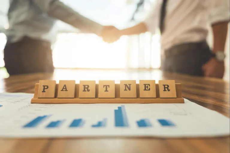 Wooden letter blocks spelling “PARTNER” on a desk with professionals shaking hands in the background, representing collaboration with an offshore software development company.