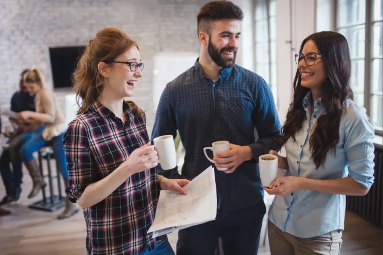 Professionals networking in a modern office, holding coffee mugs and discussing plans during a casual team meeting, showcasing offshore development collaboration and workplace communication.