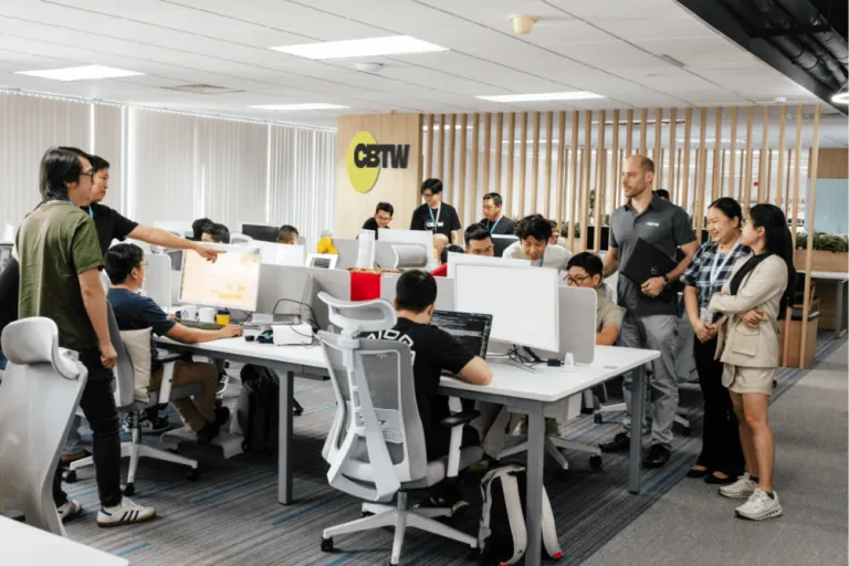 Offshore development team collaborating in a modern open-plan office, with software engineers working at computer stations while colleagues discuss project progress and technical solutions.

